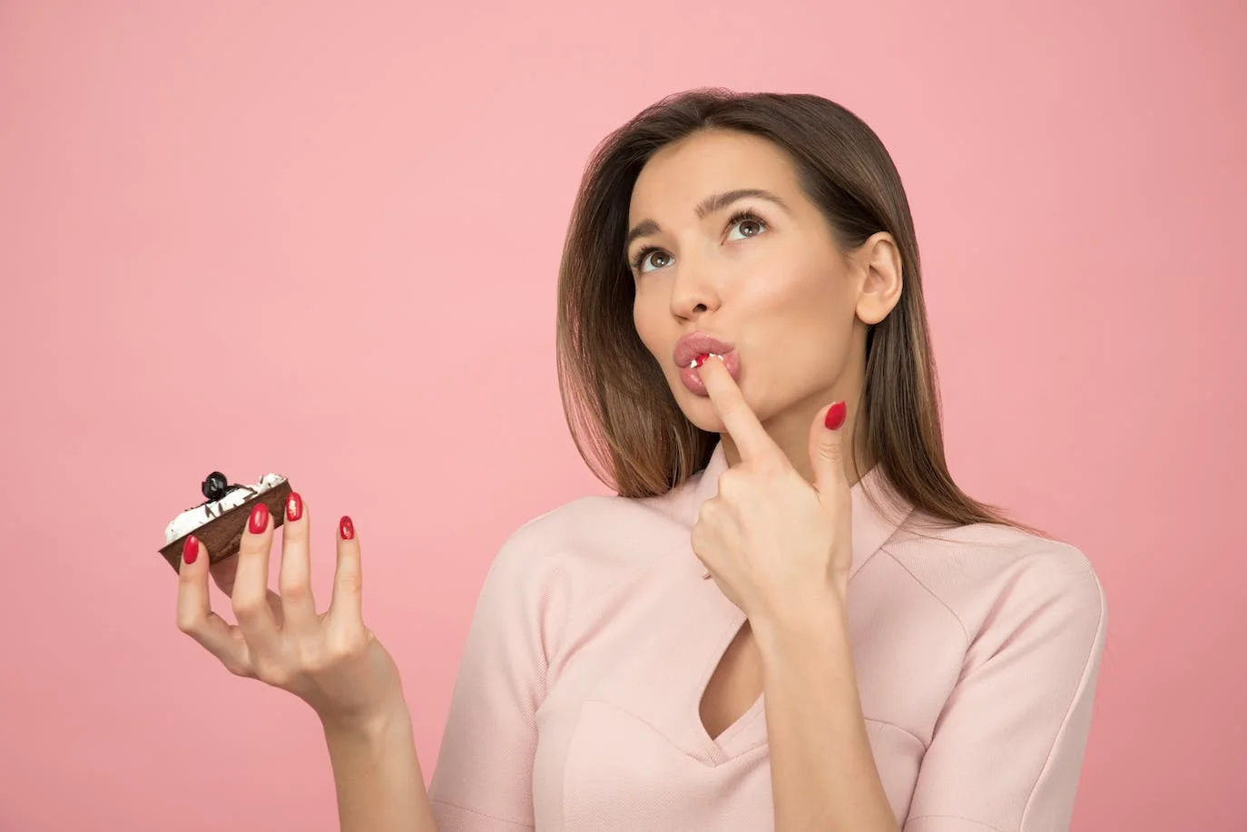 Woman photo : photography of a brown-eyed Asian woman, wearing red nail polish on both hands, holding a cupcake in her right hand and tasting the cream of the cake on one fingertip of her left hand. Plain pink background.