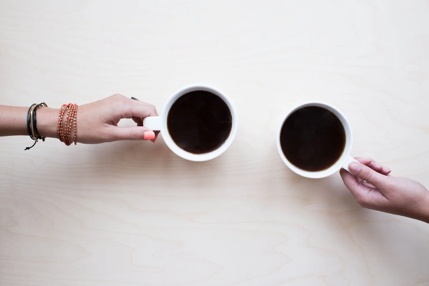 Coffee photo : top view photo of two steaming cups of coffee, each held by a hand. The one on the left is held by the hand of a white woman, of whom we can only see her hand and a part of her arm on which she wears bracelets. The one on the right is held by a white man whose hand is visible only.