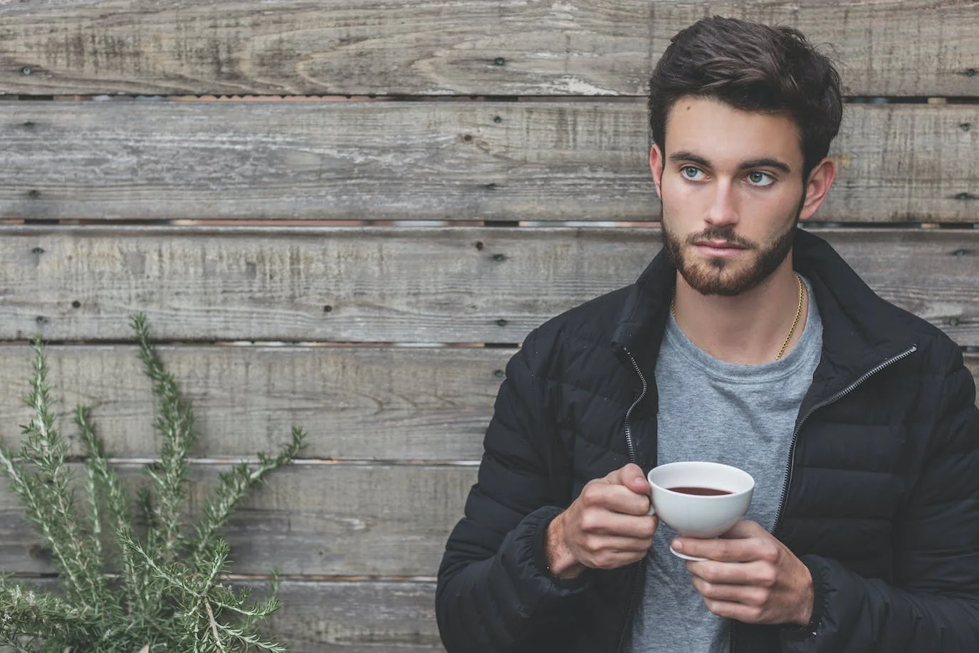 Man photo : a photography of a dark-haired man with green eyes posed in front of a gray pallet wood exterior wall, wearing a thin gold chain, a blue-gray wool sweater with a round neck, and a navy blue down jacket, holding a cup of hot chocolate with both hands.