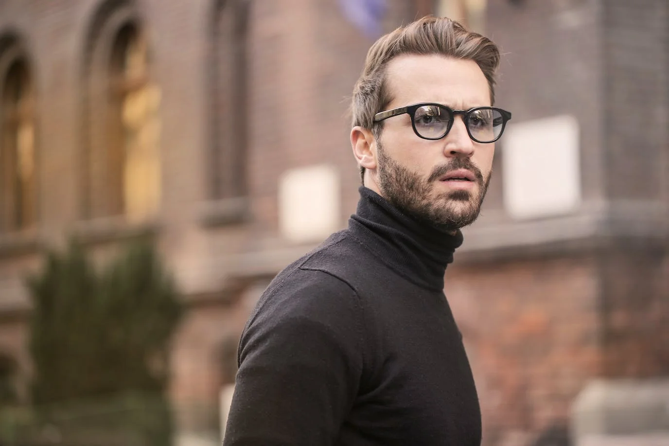 Man photo : a standing man wearing a turtleneck sweater and glasses, with a three-day beard, behind which we see a university. Outdoor photograph in daylight, blurred background.